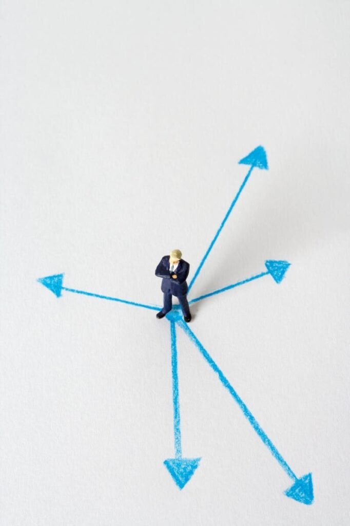 Miniature businessman stands at crossroads of blue arrows on white background, symbolizing choices.
