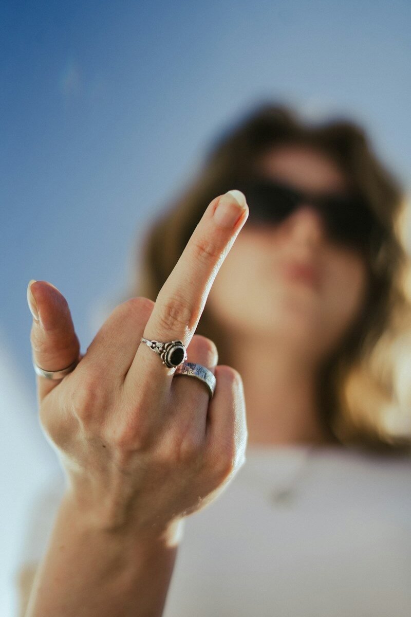 A raised middle finger in the foreground, a person in sunglasses blurred behind it